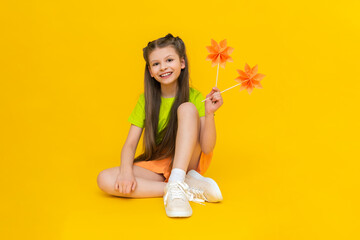 A little girl is playing with paper flowers on sticks. An origami mill. Happy kid holds paper crafts. Children's creativity.  A beautiful child is sitting on a yellow isolated background.