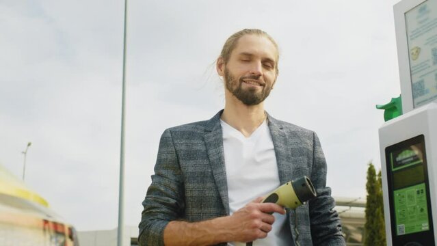 An eco urban businessman is leaning on his electic vehice dressed in a plaid suit, man use the white power cable and plug. Eco and clean energy concept. Port plugging in EV modern car