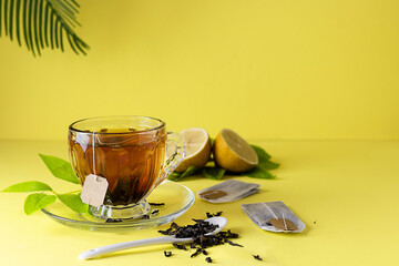 A glass cup with hot tea from a bag on a yellow table. Tea bags and lemon wedges in the background....