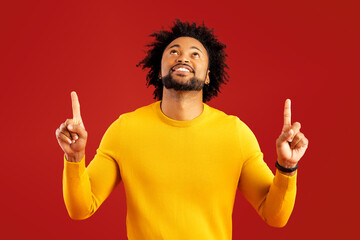 Attractive man with happy facial expression pointing up with both fingers, presenting copy space. Indoor studio shot isolated on red background