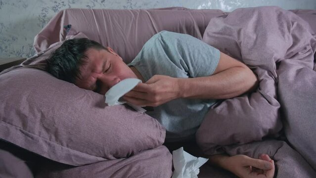 Sick man with cold, flu or virus illness with cough and running nose lying in bed and using tissues. Sneezing ill guy at home