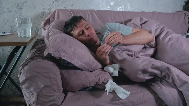 Sick man with cold, flu or virus illness drinking pill or drug for health lying in bed and using tissues. Sneezing ill guy at home