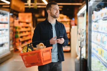Handsome man buying some healthy food and drink in modern supermarket or grocery store. Lifestyle and consumerism concept. - obrazy, fototapety, plakaty