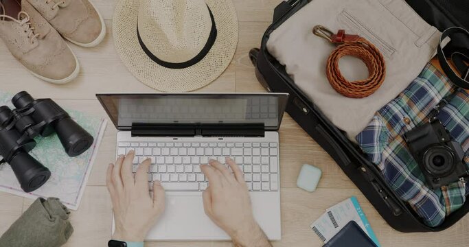 Top view of male hands using laptop typing next to packed suitcase plane tickets and map on wooden background. Modern technology and travelling concept.