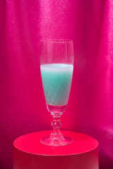 A glass of turquoise champagne wine in a crystal on a red stand. Magenta curtains background.