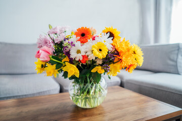 Vase with huge multicolor various flower bouquet on the coffee table with blurred background of modern cozy light living room with gray couch sofa. Giving flowers. Mother's day, birthday gift.