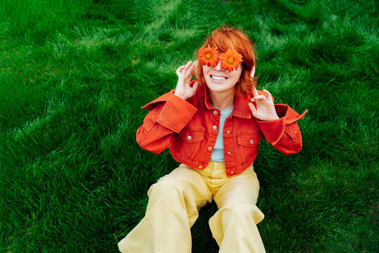 Happy Smiling Redhead Woman With Freckles In Red Orange Gerbera Flower Glasses Sitting On Green Grass And Listening To Music In Wireless Headset. Positive Emotions. Fashion. Spring, Summer Mood