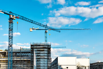 Two construction tower cranes at a construction site against a blue sky with clouds. Modern high-rise office building under construction. Copyspace top right, horizontal © Iuliia