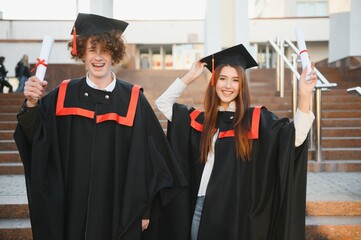 Portrait of happy graduates. Two friends in graduation caps and gowns standing outside university...