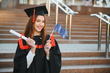 Happy cute brunette caucasian grad girl is smiling. She is in a black mortar board, with red tassel, in gown, with nice brown curly hair, diploma in hand. - obrazy, fototapety, plakaty