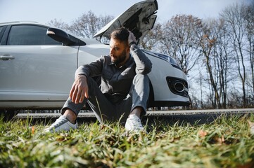 Frustrated sad upset guy driver is sitting near a broken car after road accident, crash. Young...