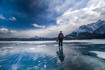 young man with beard in sweater walks on blue ice of abraham lake and holds in hands ice skates in jasper and banff national park - obrazy, fototapety, plakaty