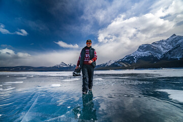 young man with beard in sweater walks on blue ice of abraham lake and holds in hands ice skates in jasper and banff national park - obrazy, fototapety, plakaty