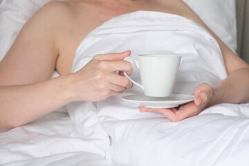Fototapeta na wymiar Woman in bed holds a white cup on a saucer in her hands