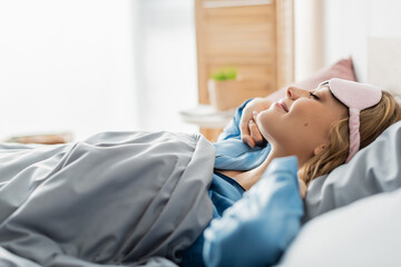 side view of pleased woman in sleeping mask lying under blanket in comfortable bed.