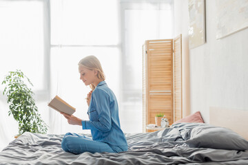 carefree young woman in blue silk nightwear reading book in modern apartment.