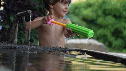 One happy child spraying water with foam toy blaster at outdoor backyard in summer day. Kid playing with water shirtless. Happiness concept