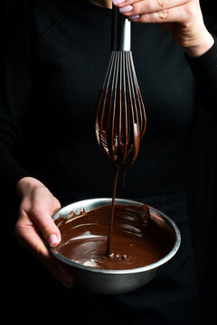 A bowl of melted chocolate and a kitchen whisk are held by hands. Kitchen utensils. On a black background.