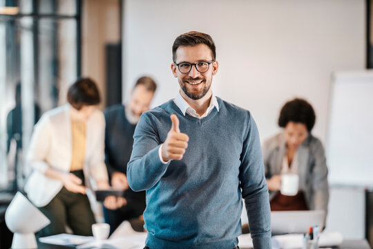 Portrait of a happy businessman giving thumbs up at the camera while his colleagues working in a blurry background at the office.