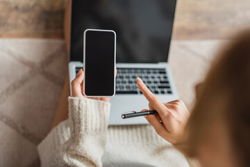 partial view of blurred freelancer pointing with finger at smartphone with blank screen near laptop.