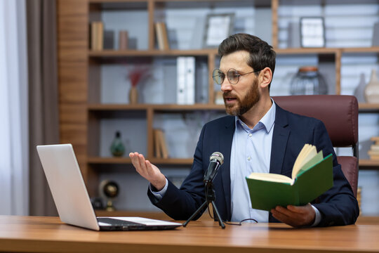 Online Presentation Of The Book. A Young Man Writer, Author In A Suit Sits In The Office At A Table In Front Of A Camera, A Laptop And A Microphone, Holds A Book, Reads To Listeners On A Video Call.