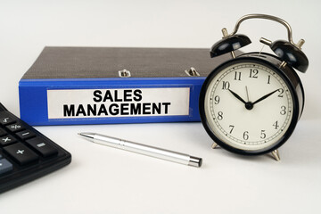 On a white surface, an alarm clock calculator and a folder with the inscription - Sales Management