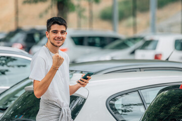 young man with mobile phone looking at cars with expression of success