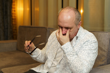 A mature man holds glasses with his hand, the other rubs his eyes.