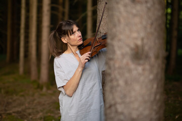 Pretty woman is simply dressed in nature in forest playing violin