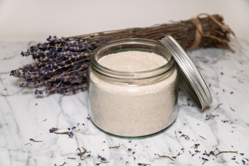 Close up of natural handmade bath salt in transparent glass jar with aluminium lid on white marble background with dry lavender flowers.