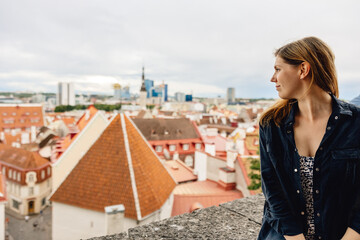 woman sits on wall overlooking old European town centre, with view of medieval colourful buildings in background, Tallinn, Estonia