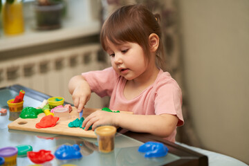 The little girl is carving plasticine figures out of the house at the table.