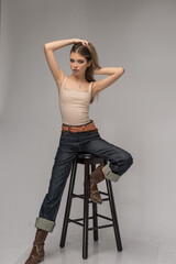 Attractive girl in blue jeans and long legs posing challenging in front of a background. Classic fashion scene. Indoor glamour photo. Sensual young  woman with long  hair and brown boots on high chair