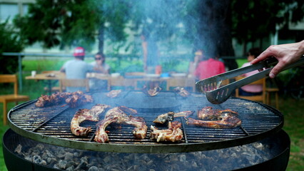 Person preparing meat on grill outside with friends and family in background. Closeup hand holding clamp cooking food at BBQ party
