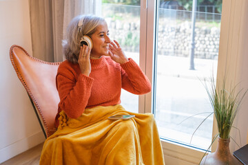 Senior woman with white hair sitting on the sofa at home listening to music and relaxing in the living room. Concept: technology, headphones, relax