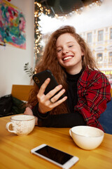 Close-up portrait of happy young woman making selfie on mobile phone sitting at table with laptop by window in cafe. Pretty redhead Caucasian lady having leisure activity in coffee shop.