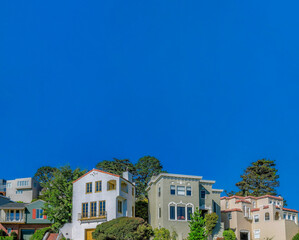 San Francisco hill houses with beautiful architecture seen from the street. Houses surrounded by lush trees against blue sky on a sunny day at a California neighborhood.