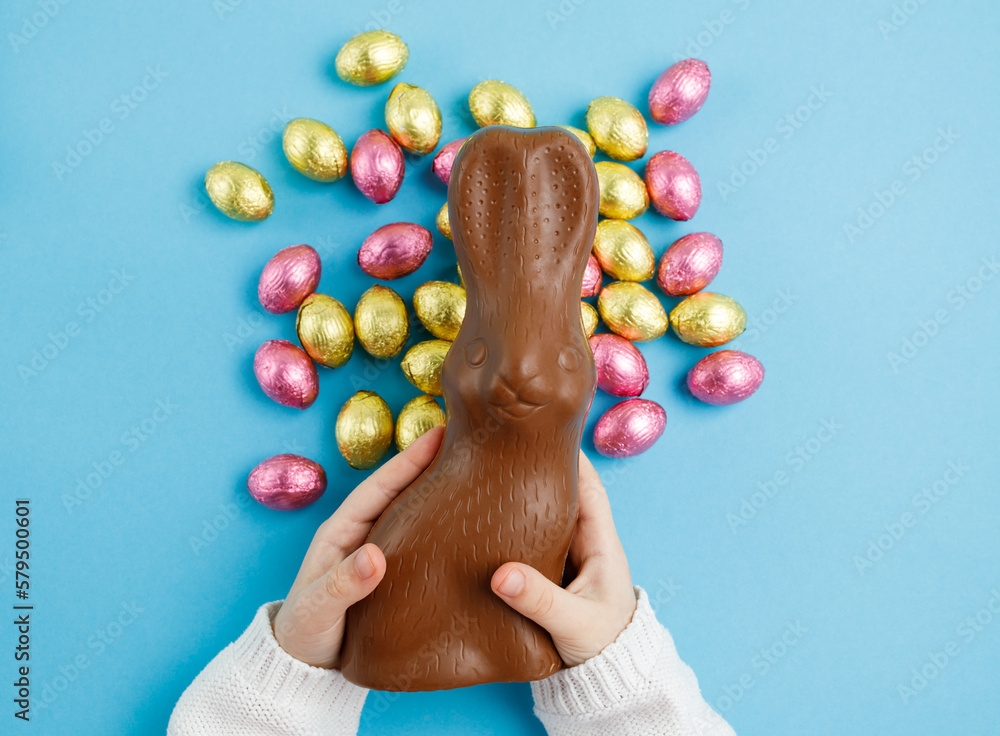 Wall mural child's hands holding easter chocolate bunny on blue background with pink and golden candy eggs, eas