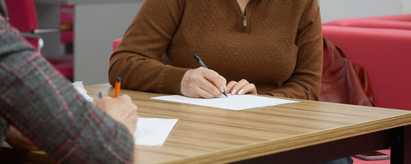 Two women, sitting opposite each other at a wooden table, fill out documents, forms, contracts, tests, applications or resumes. Selective focus.