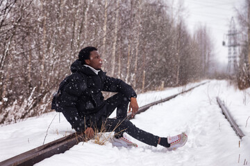Fototapeta na wymiar Pensive african american man sitting on railway track in snowy russian winter at countryside; looking away. Sad black guy posing outside; falling snow. Lifestyle activity concept. Copy text space