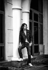 Black and white photo of a young man with long hair.in a leather jacket and jeans