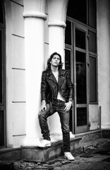 Black and white photo of a young man with long hair.in a leather jacket and jeans