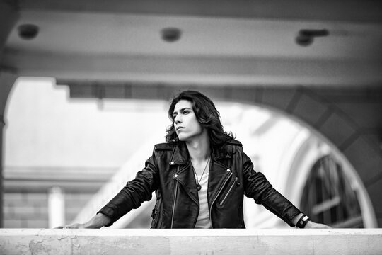 Black and white photo of a young man with long hair.in a leather jacket and jeans