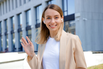 Portrait of the smiling positive caucasian woman standing waving hello to the camera. Smiling female worker wearing earphones looking at the camera standing outside of the office building.