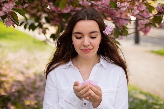 Young woman romantic close up portret in a blooming sakura garden. Wind is hairs. Spring story. Brown-haired girl smiles with a flower in her hairs closing her eyes and enjoy of spring mood
