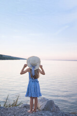 A girl in a hat with long blond hair admires the sunset on the shore.