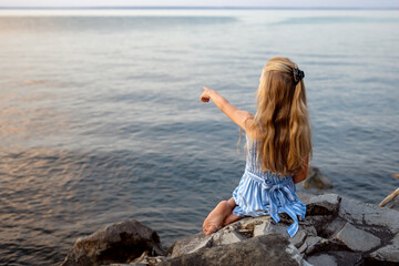 Beautiful girl with long blond hair sits on a stone on the banks of a river, lake, sea.