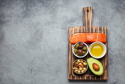 Food sources of healthy unsaturated fat and omega 3: fresh raw salmon fillet, avocado, olives, nuts on cutting board, rustic stone background top view. Healthy nutrition and keto diet, space for text