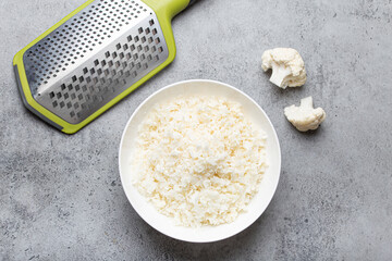 Raw cauliflower rice or couscous in a white bowl with grater, healthy low carbohydrates vegetable side dish for keto diet and healthy low calories nutrition, grey concrete rustic background