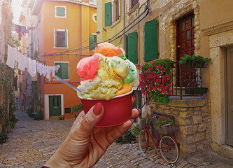 Woman hand hold  beautiful bright sweet ice - cream cone with different flavors  held in hand on the background of old street  in  Rovinj .Rovinj is a tourist destination on Adriatic coast of Croatia - 579510875